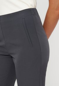 Gros plan d'une personne portant un pantalon ajusté gris foncé avec une poche latérale et une taille élastique, associé à un haut blanc.