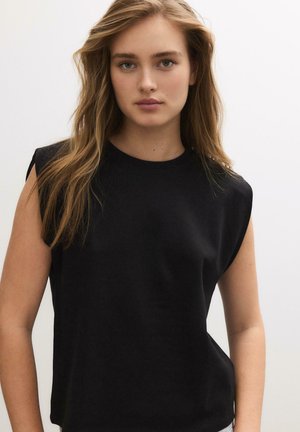 Jeune femme aux longs cheveux châtain clair portant un haut noir sans manches, face à l'avant sur un fond clair uni.