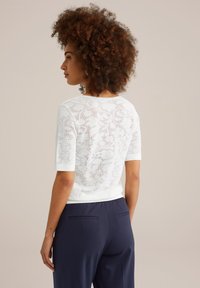Haut en dentelle blanche à manches courtes, avec un motif floral et un col rond, associé à un pantalon bleu marine foncé. Texture lisse.