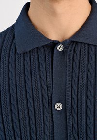 Granatowy, dzianinowy polo z teksturowanym wzorem w warkocz oraz klasycznym kołnierzykiem, z jednym okrągłym srebrnym guzikiem z przodu.