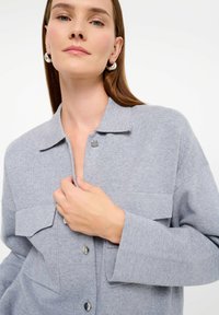 Svetlo šedá pletená košeľa s golierom, dvomi náprsníkovými vreckami a zaoblenými gombíkmi. Mäkka textúra, uvoľnený strih, dlhé rukávy.