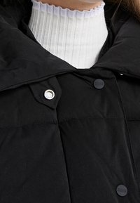 Chaqueta de plumas negra con cuello alto. Presenta un botón de presión metálico y una tela texturizada. Se lleva sobre un suéter de cuello alto de canalé blanco.