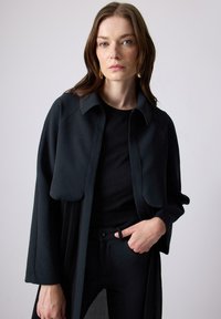 Black long coat with a collar, featuring a pleated design at the front. Worn over a black top, paired with black pants. Smooth texture.