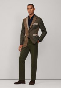 Bruine en groene geruite blazer met contrasterende tan zakken, gecombineerd met een marineblauw shirt en olijfkleurige broek. Bruine schoenen maken de look compleet.