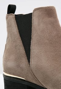 Bottine en daim taupe avec panneau élastique latéral noir et languette au talon, présentant un détail de semelle noir et beige.