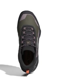 Scarpa da trail con una parte superiore verde scuro e grigia, superficie testurizzata, accenti viola, suola in gomma e un logo riflettente. Design con chiusura a lacci.