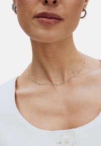 Srebrny naszyjnik z równomiernie rozmieszczonymi małymi białymi koralikami, noszony blisko szyi. Modelka ma na sobie teksturowany biały top z guzikiem.