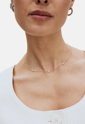 Sudraba kaklarota ar mazām baltām pērlītēm, kas vienmērīgi izvietotas un valkāta cieši pie kakla. Modelis valkā baltu teksturētu augšu ar pogu detaļu.