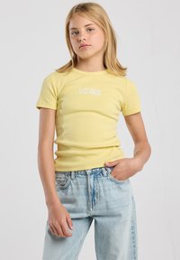 Keltainen ribattu t-paita, jossa on pyöreä kaula-aukko ja lyhyet hihat, jossa on valkoinen graafinen printti rinnassa. Yhdistetty vaaleisiin farkkuihin.