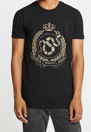 Person trägt ein schwarzes T-Shirt mit einem goldenen Schlangenemblem, einer Krone, einem Lorbeerkranz und dem Text "Glorious Gangsta MCMLXX" auf der Vorderseite.