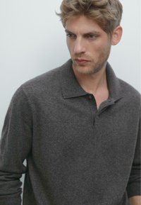 Dunkelgraues, langärmliges Poloshirt aus weichem Stoff, mit einem Kragen und einer Knopfleiste mit zwei Knöpfen, getragen von einem Model vor einem hellen Hintergrund.