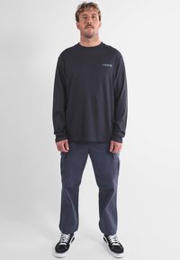 T-shirt bleu marine à manches longues avec logo sur la poitrine, associé à un pantalon cargo gris avec poches latérales et des baskets noires à semelles blanches.