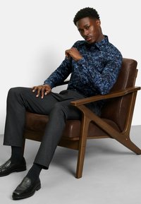 Hellblau gemusterte Bluse mit Blumenmotiven aus Stoff, kombiniert mit dunkelgrauen strukturierten Hosen, schwarzen Loafers, sitzend auf einem Holzstuhl.