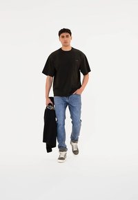 Fekete, oversized T-shirt diszkrét logóval, világos kék farmerral és szürke-fehér sportcipővel párosítva, fekete dzsekit tartva.