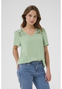 Chemisier vert menthe avec des détails en dentelle sur les épaules, manches courtes et décolleté en V, associé à un jean en denim bleu.