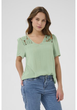 Chemisier vert menthe avec des détails en dentelle sur les épaules, manches courtes et décolleté en V, associé à un jean en denim bleu.