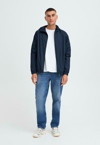 Navy windbreaker with hood, lightweight fabric, elastic cuffs, front zipper, worn over a white t-shirt and blue jeans, featuring white sneakers.