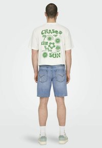 La camiseta crema presenta un texto gráfico en verde y iconos en la parte posterior; combinada con pantalones cortos de mezclilla azul claro y zapatillas de color claro.