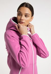 Roze hoodie van zacht materiaal, met een hoge kraag, witte rits en een comfortabele capuchon. Het ontwerp benadrukt comfort en pasvorm.