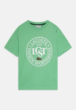 Zielona bawełniana koszulka z okrągłym białym graficznym akcentem podkreślającym nazwę marki "LACOSTE" oraz logo krokodyla na dole.