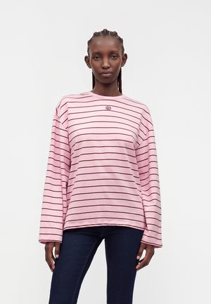 Femme aux cheveux tressés portant une chemise rose à manches longues avec de fines rayures horizontales rouges et un pantalon bleu foncé, debout devant un fond uni.