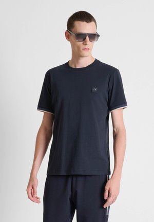 Giovane uomo con occhiali da sole scuri, t-shirt nera a maniche corte e pantaloni neri abbinati, in piedi contro uno sfondo chiaro e semplice.