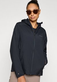 Casaco leve preto com fecho de correr e capuz, com punhos elásticos e detalhe de logótipo. Usado por cima de um top preto, combinado com óculos de sol castanhos.