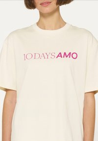 Lys beige kortærmet t-shirt med rund hals, prydet med et lilla grafisk print, der står "10 DAYS AMO" på forsiden.