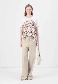 Biely tričko s leopardím vzorom vpredu; béžové široké nohavice; modelka drží štruktúrovanú svetlobéžovú kabelku; jednoduché pozadie.