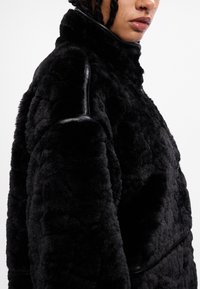 Chaqueta de pelo sintético negro con un alto cuello, que presenta una textura acolchada y detalles de cuero en los hombros y las costuras.