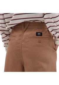 Brązowe spodnie o gładkiej fakturze, z dwiema tylnymi kieszeniami, zapięciami na guziki oraz czarną metką marki, w połączeniu z koszulą w paski.