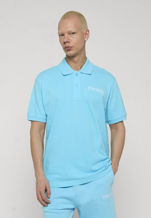 Person mit kurzen blonden Haaren, trägt hellblaues Polohemd und passende Hose mit "bruno banani"-Logo, posiert vor neutralem Hintergrund.