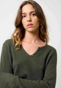 Olivengrøn sweater med V-udskæring, der har tre knapper nær halsen, lange ærmer og blødt, struktureret stof.