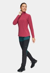 Polarowa bluza z połową zamka w kolorze burgundowym, zestawiona z turkusowymi i czarnymi spodniami. Czarne buty trekkingowe z teksturowaną podeszwą. Minimalne detale z logo.