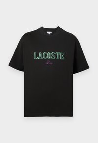 T-shirt preta de algodão com mangas curtas, com texto verde "LACOSTE" e "Paris" em roxo abaixo. Design simples, decote redondo padrão.