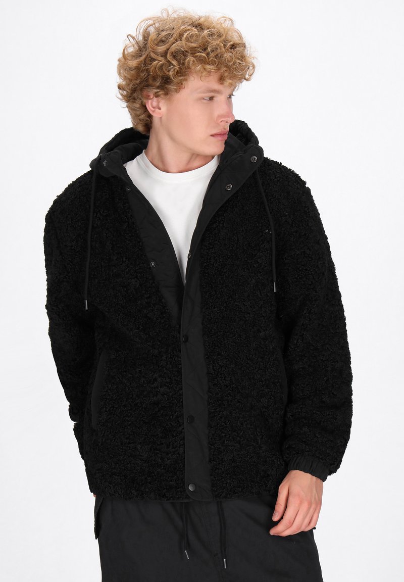 Svart fleecejacka med en texturerad, fluffig yta, kontrasterande svart insida, dragkedja i huvan och tryckknappsstängning. Långa ärmar och elastiska muddar.