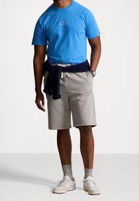 Blue cotton t-shirt with embroidered logo, gray fleece shorts, navy sweater tied at waist, beige sneakers with white accents, ribbed gray socks.