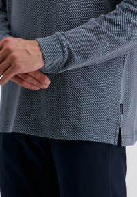 Langärmliges, navyblaues Hemd mit strukturiertem Muster, das hellblaue und weiße Punkte aufweist, seitliche Schlitze und ein kleines Markenetikett am unteren Saum hat.