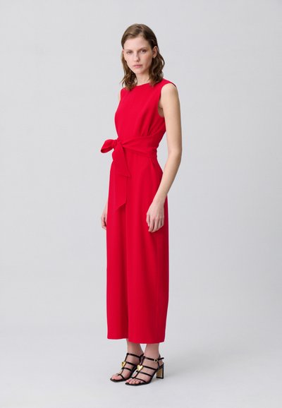 Mono rojo sin mangas con detalle de cintura atada y patas anchas, hecho de una tela suave. Lo lleva con tacones negros con correas y acentos dorados.