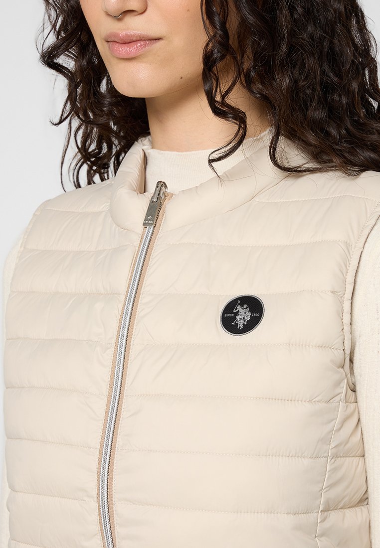 Beige gesteppte Weste mit hohem Kragen und Reißverschluss. Verfügt über horizontale Steppungen und ein schwarzes Logo-Patch auf der linken Brustseite.