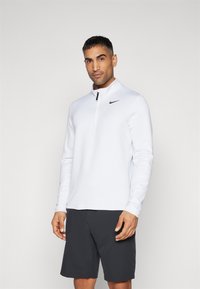 Cămașă albă cu mâneci lungi, cu fermoar, dintr-un material texturat, având un logo Nike negru pe piept. Asortată cu pantaloni scurți închisi la culoare.