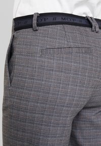Grå mönstrade byxor med houndstooth-design, med ett svart midjeband med loggtext och detaljerad bakficka.