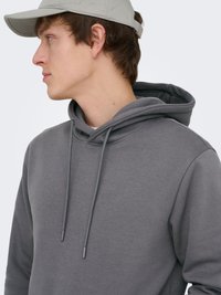 Jonge man met een grijze hoodie en een lichtgrijze baseballpet die naar links kijkt tegen een witte achtergrond.