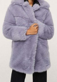 Persona che indossa un morbido e soffice cappotto di pelliccia sintetica lavanda, mentre buttona la parte frontale, abbinato a pantaloni neri su uno sfondo neutro.