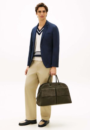 Joven con blazer azul marino, suéter blanco de cuello en V, pantalones beige, mocasines negros, sosteniendo un bolso grande de ante oscuro por las asas.