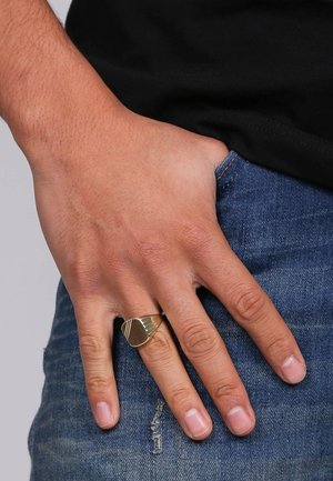 Goldener Ring mit geometrischem Design und Rillen, getragen an einer Hand, die auf Jeans ruht. Die Hand zeigt kein sichtbares Schmuckstück außer dem Ring.