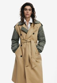 Trenchcoat met een tan basis, groene mouwen, een dubbelknopen ontwerp, zwarte knopen en een riem. Gladde textuur met grote zakken.