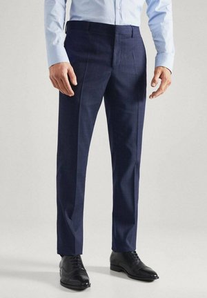 Mężczyzna ubrany w granatowe eleganckie spodnie, czarne buty wizytowe oraz jasnoniebieską koszulę z długim rękawem, stojący na tle jednolitego jasnego tła.