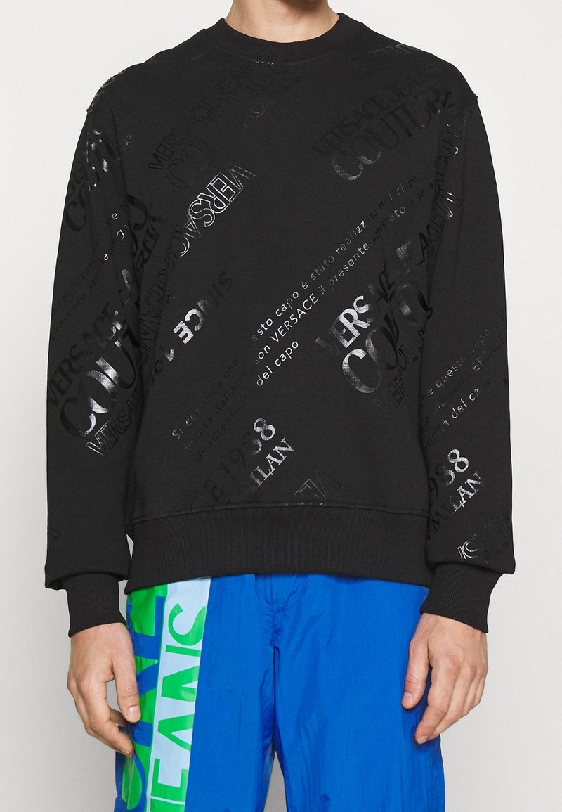 Personne portant un sweat-shirt noir Versace Couture avec un texte argenté et un pantalon bleu avec un imprimé vert et blanc.