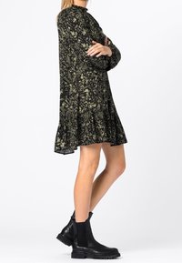 Vestido negro con patrón abstracto verde, con un dobladillo a la altura de la rodilla, corte holgado y tejido texturizado. Usado con botines negros hasta el tobillo que tienen lados elásticos.